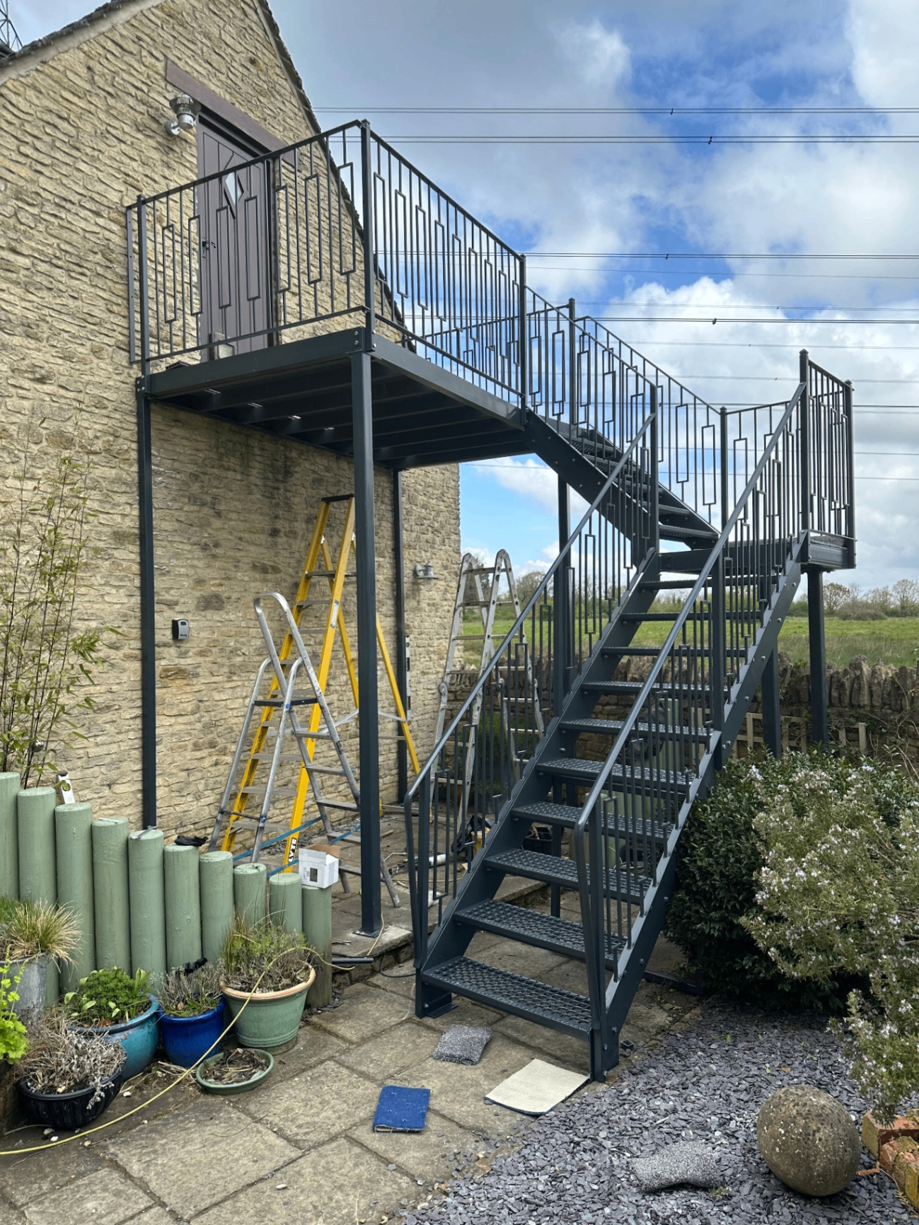 External steel staircase and landing with decorative railings against a cotswold-stone building.